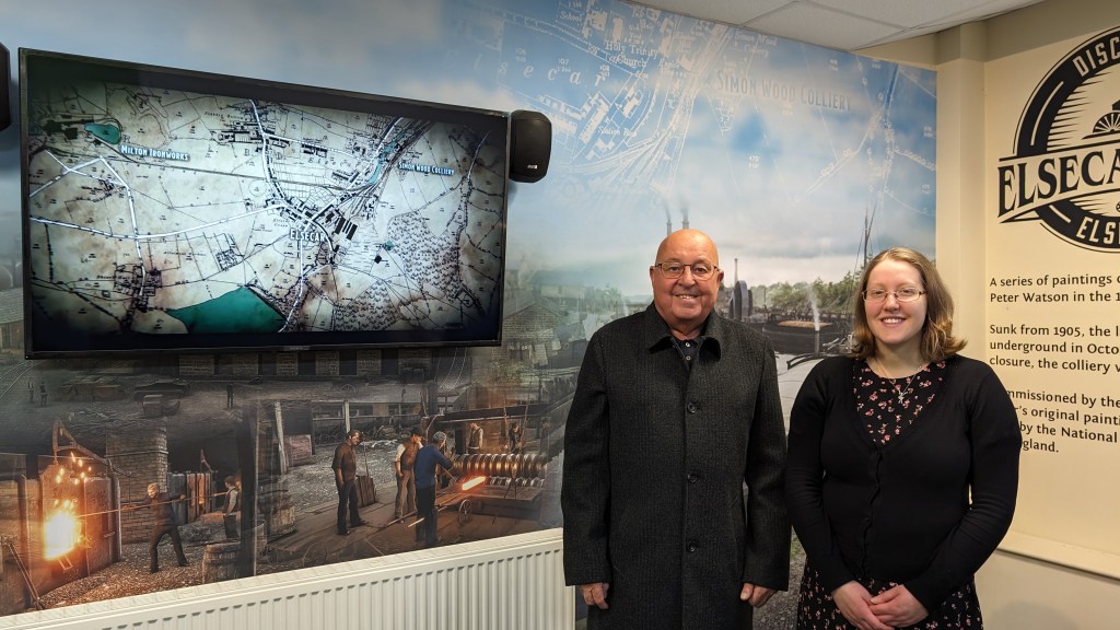 Two individuals are standing indoors in front of a wall with historical maps and imagery from an industrial era displayed. The wall includes the name "Elsecar." One person is wearing a black coat, and the other is in a black dress with floral patterns. Both are smiling.