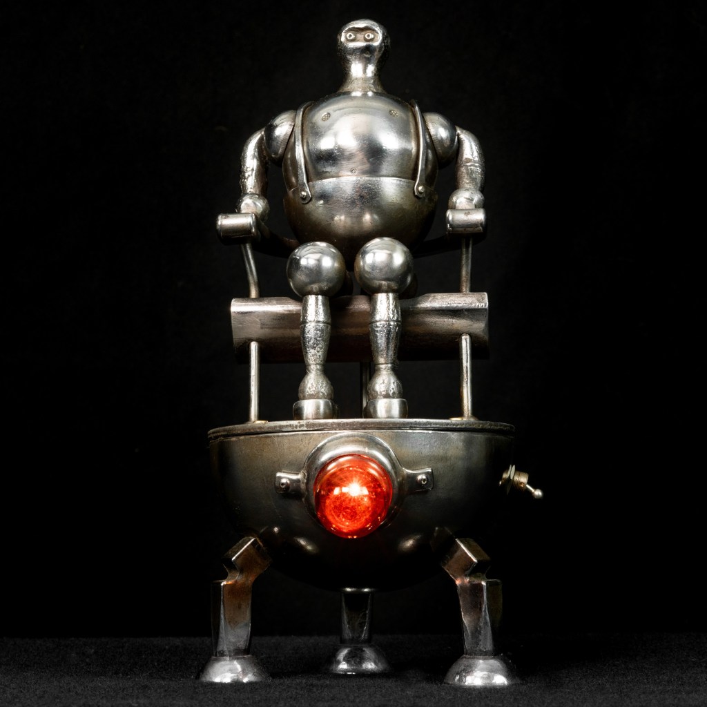 A metallic robot with a round body and articulated arms and legs sits atop a spherical, four-legged base. It features a bright red light at the center of the base, creating a futuristic appearance against a dark background.