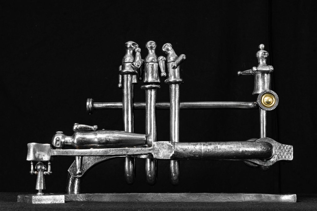 A black and white photo of a metallic sculpture featuring stylized figures on a platform. Some figures stand upright, while one lies horizontally. The artwork appears abstract, composed of rods and rounded shapes on a dark background.