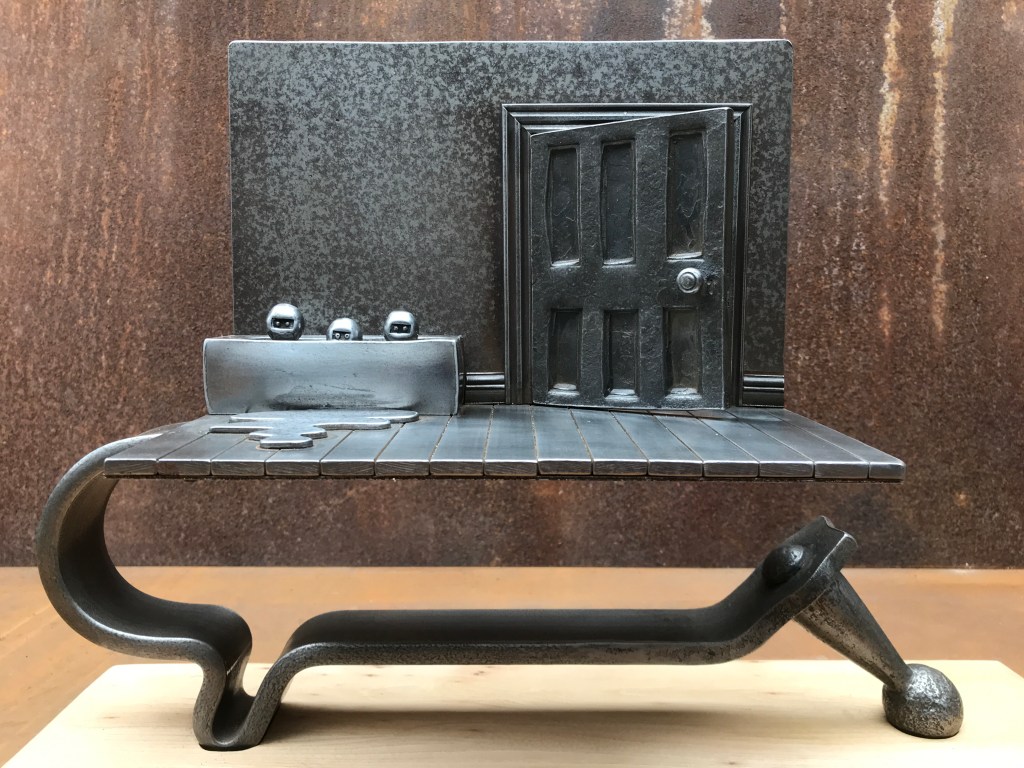 Metal sculpture depicting a table with a textured surface, featuring an open door and three round objects next to a rectangular sink on the table. The background is a rusty brown wall.