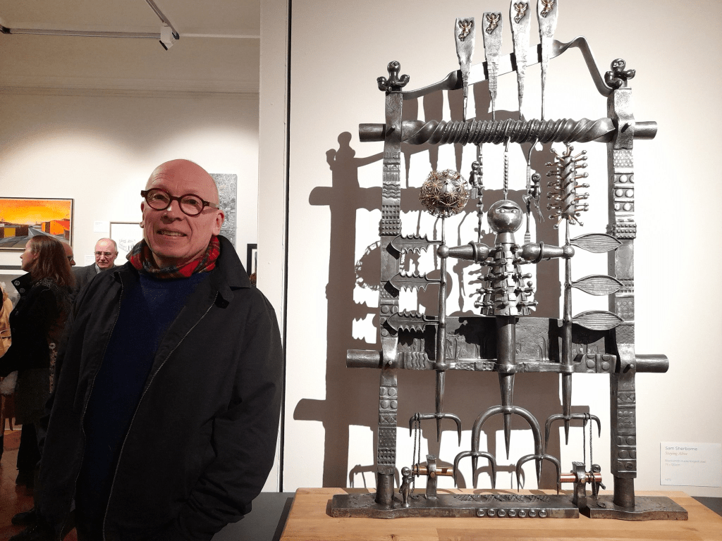 A person wearing glasses and a scarf stands next to a large, intricate metal sculpture on display in a gallery. Other artworks and people are visible in the background.