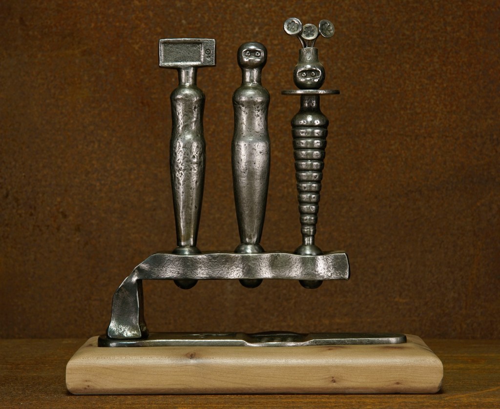 A metal sculpture featuring three abstract, humanoid figures on a wooden base. Each figure has a distinct head shape: a rectangular block, a stylized face, and a rounded drilled cone with circular appendages. The background is textured brown.