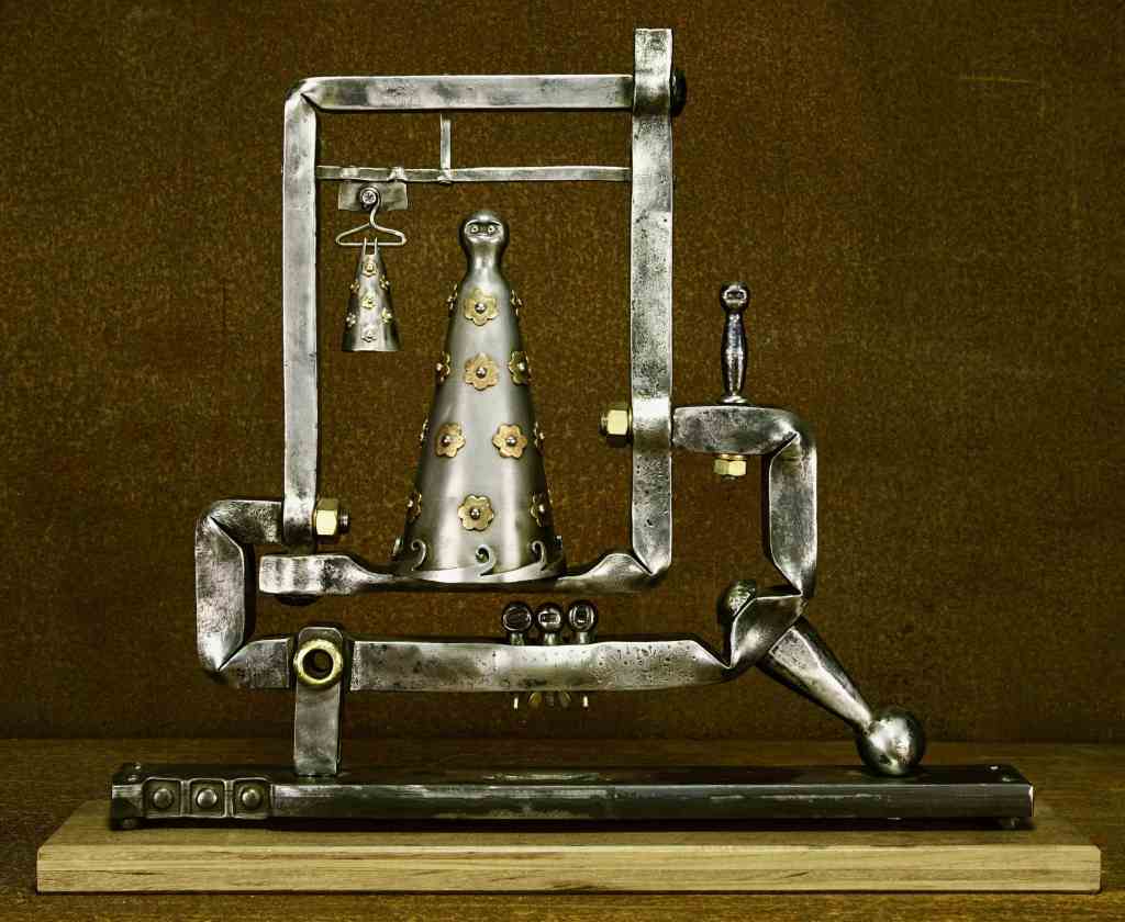 Metal sculpture of two abstract human figures with outstretched arms, perched atop a mechanical-looking base. The figures have geometric patterns and hold small objects, set against a brown background on a wooden platform.