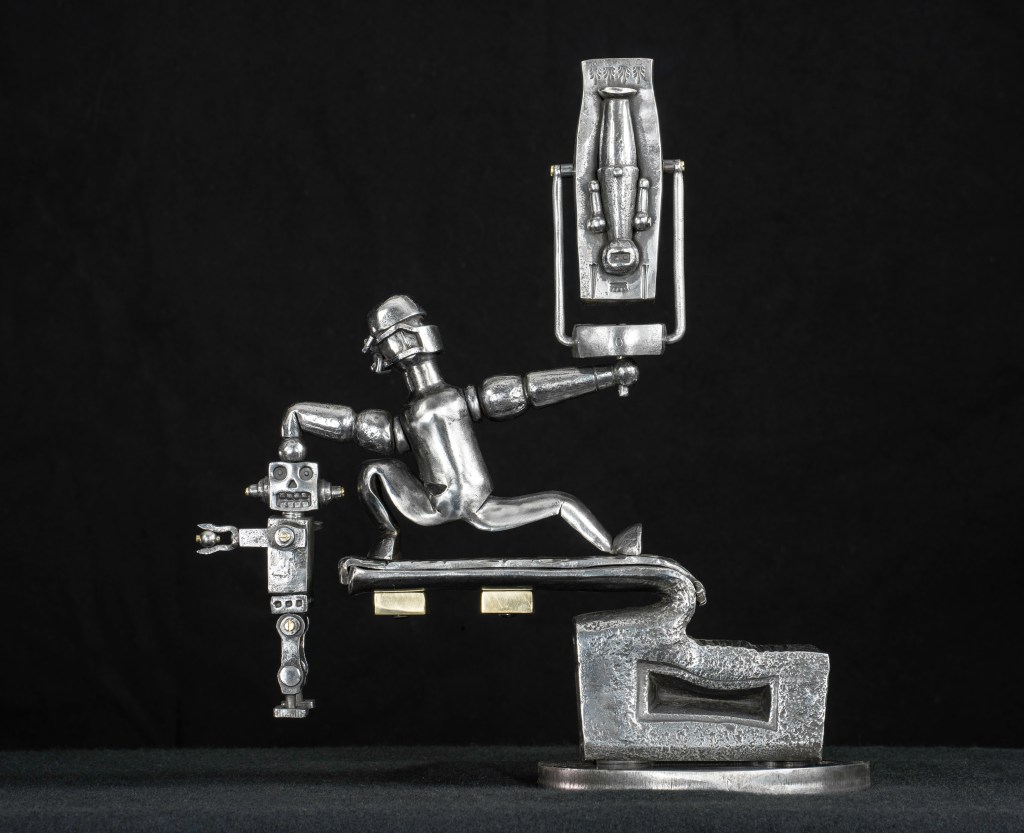 A metal sculpture featuring three abstract, humanoid figures on a wooden base. Each figure has a distinct head shape: a rectangular block, a stylized face, and a rounded drilled cone with circular appendages. The background is textured brown.