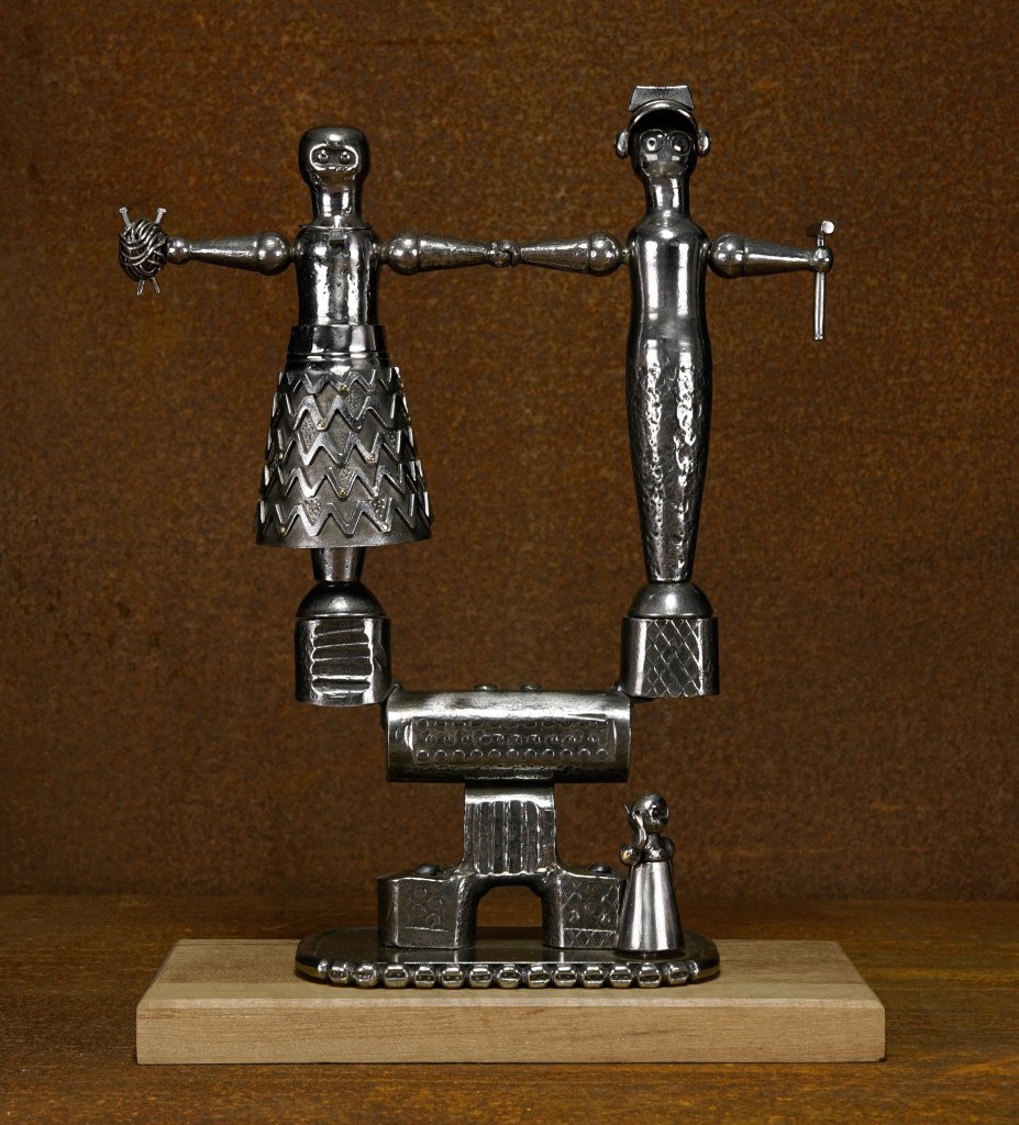 Metal sculpture of two abstract human figures with outstretched arms, perched atop a mechanical-looking base. The figures have geometric patterns and hold small objects, set against a brown background on a wooden platform.