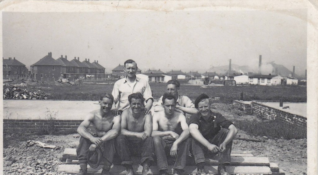 Six men are posing on a stack of wooden planks at a construction site. Four are shirtless, and two are wearing shirts. A row of houses and several smokestacks are visible in the background. The photo has an old, sepia tone.