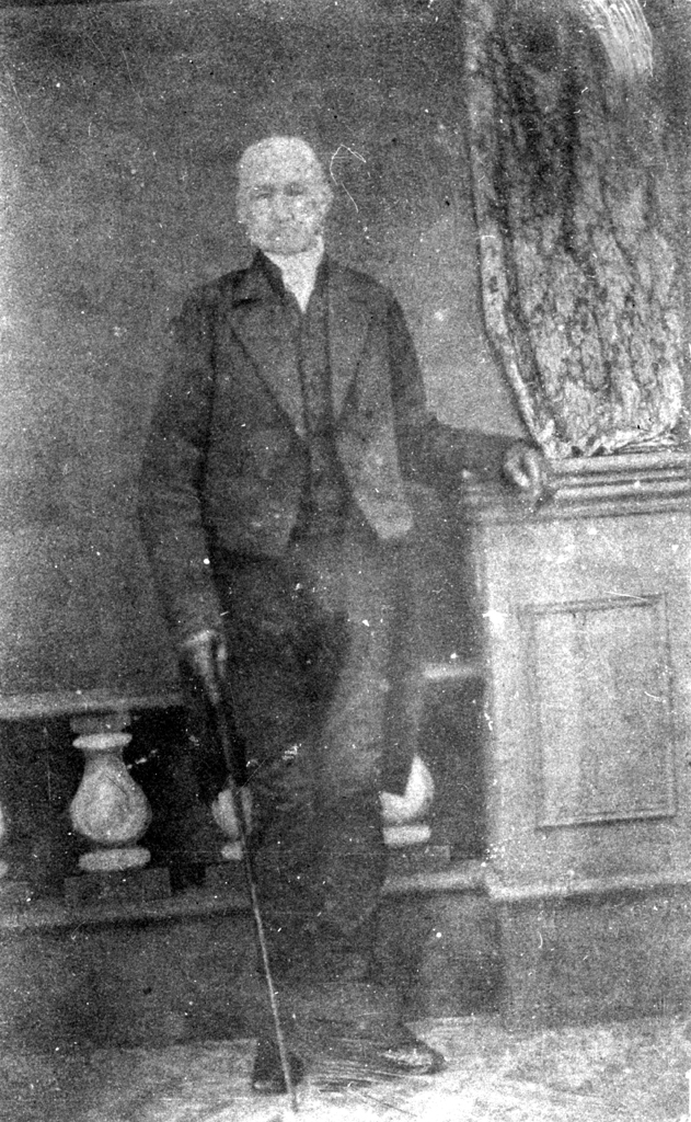 A sepia-toned, vintage photograph of an elderly man in a suit standing with a cane. He is leaning against a wooden cabinet with draped fabric. The background is plain, enhancing the historical feel of the image.
