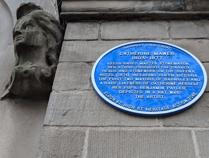 A blue historical plaque on a stone wall honors Catherine Mawer, a Leeds-based master stonemason, highlighting her work on the Queens Hotel in 1874. The plaque is accompanied by a stone carving of a face.