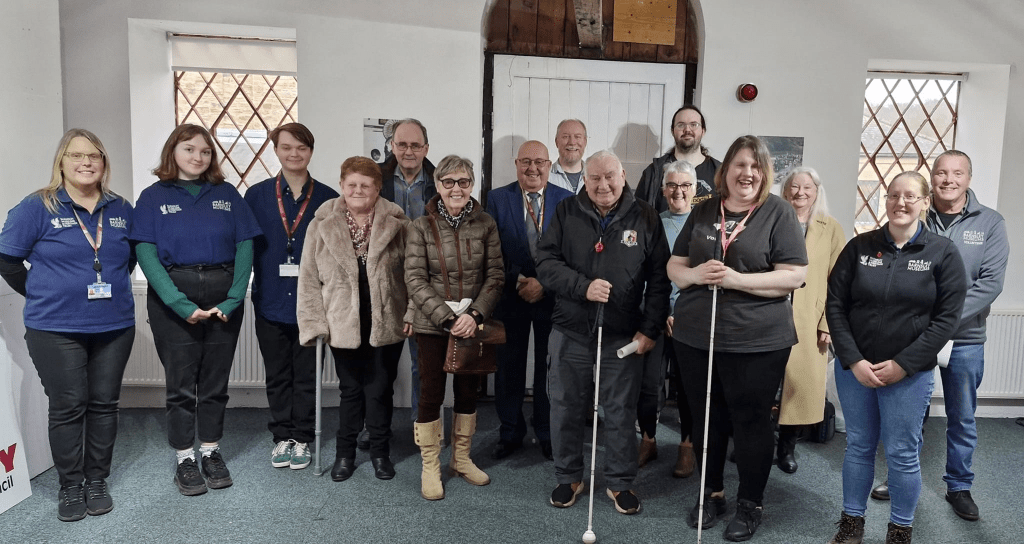 A group of people are standing together indoors. Some are using canes. They are smiling and dressed in casual clothing. The background features a white wall with lattice windows and a wooden door.