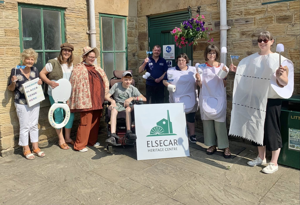 A group of people stand outside the Elsecar Heritage Centre holding toilet brushes and seats. They are dressed in various costumes, including historical attire and a toilet paper outfit, smiling and posing with a sign for the heritage center.
