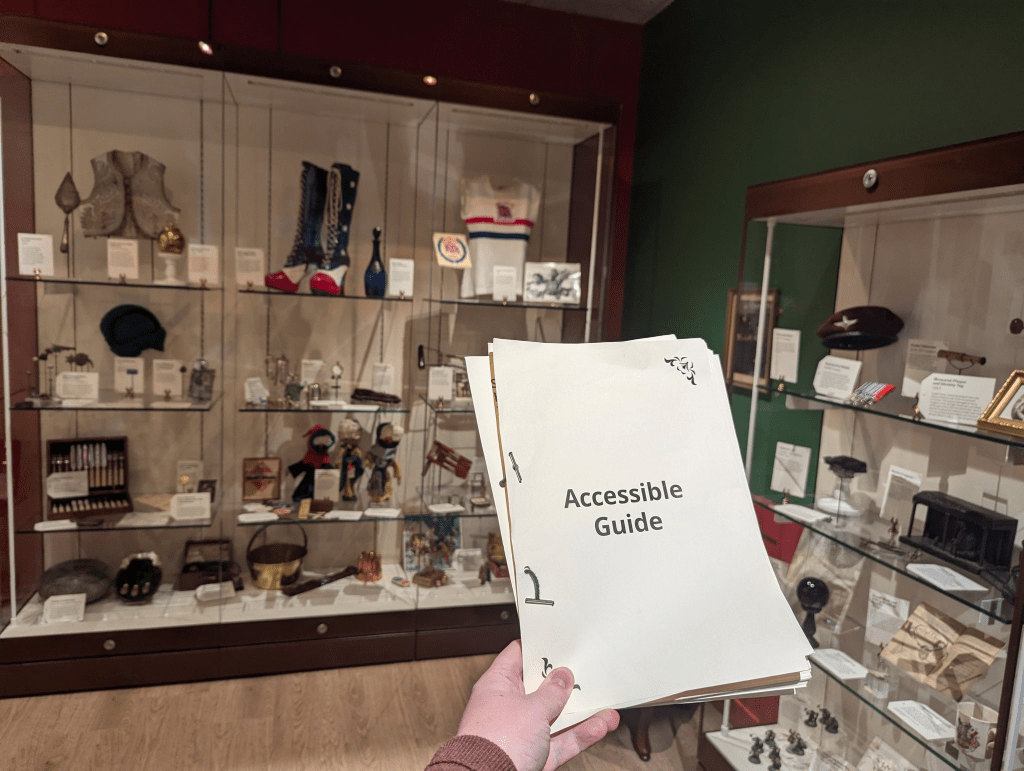 A person holds an "Accessible Guide" in front of museum exhibit cases filled with various artifacts, including hats, bottles, and decorative items. The display cases are well-lit and arranged neatly against the wall.