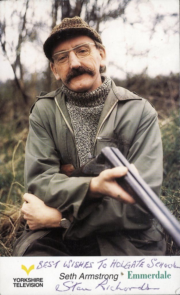 A man with glasses and a mustache holds a shotgun, wearing a knitted sweater and a green jacket, posing outdoors with trees in the background. The image includes a handwritten note with best wishes to Holgate School from Stan Richards.