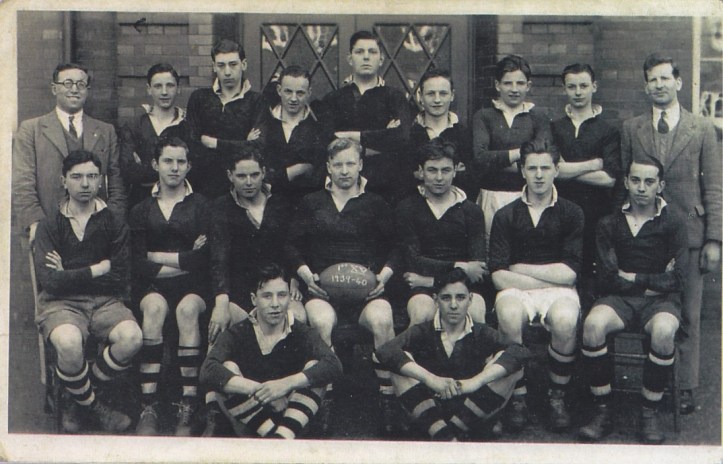 A vintage black-and-white photo of a rugby team posed in front of a brick building. The players, dressed in dark jerseys and striped socks, sit and stand in rows with two men in suits at the ends. One player holds a rugby ball.