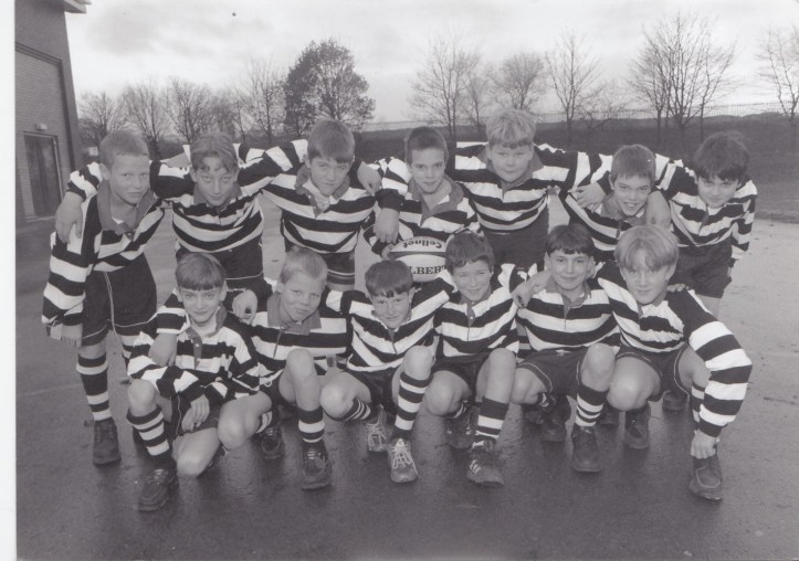 A black and white photo of a young boys' rugby team. They are wearing striped jerseys and shorts, posing together in two rows outdoors on a cloudy day, with trees in the background.