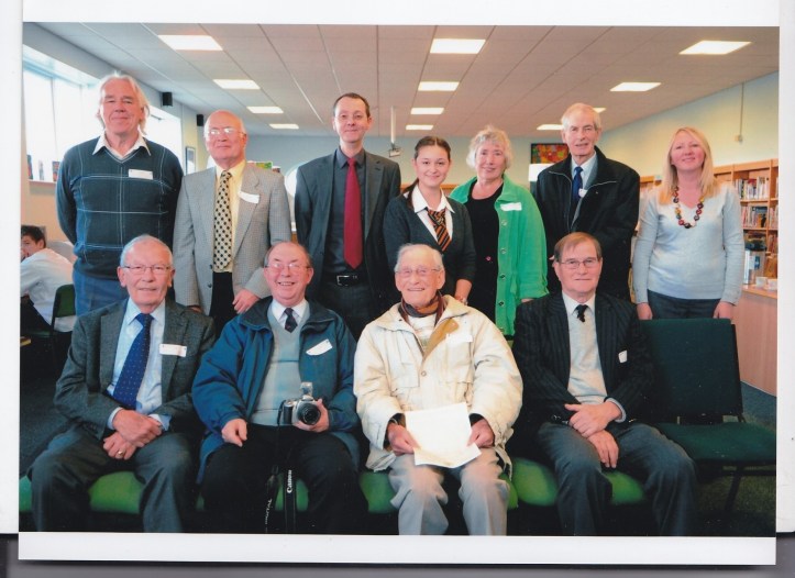 A group of eleven people posing for a group photo indoors. Four men are seated in the front row, and seven people, including women and men, are standing behind them. Some individuals are wearing name tags. They are in a room with shelves and large windows.