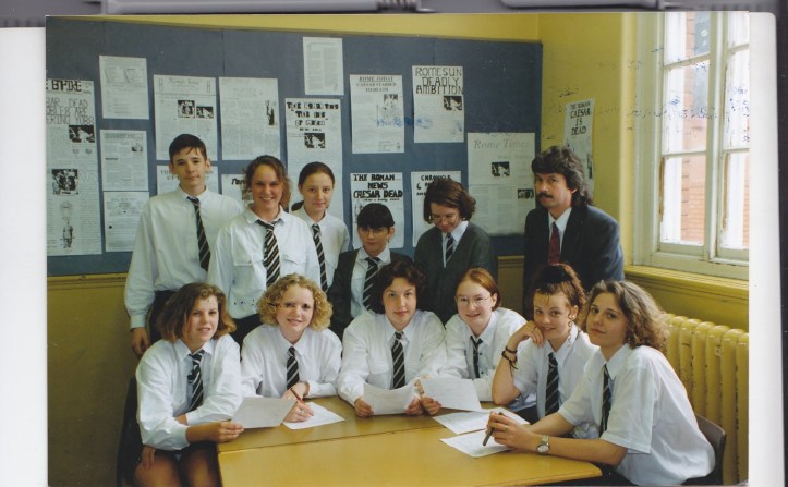 A group of students in uniforms and a teacher pose around a table in a classroom. They are smiling, with some holding papers. Behind them, a bulletin board displays various notices and articles.