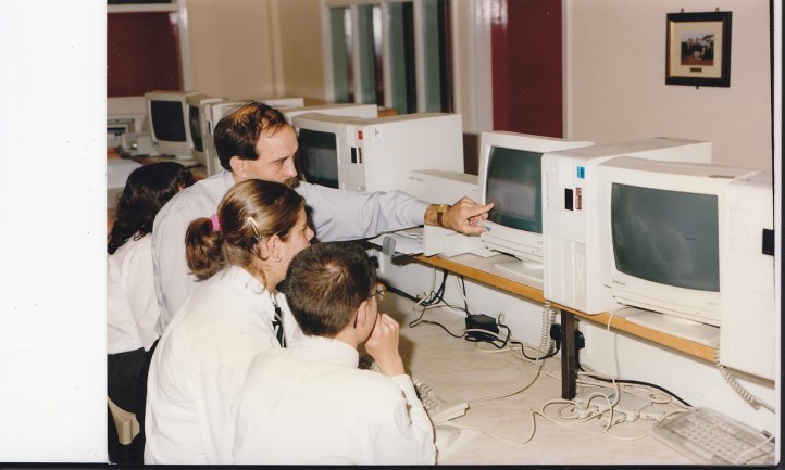 A group of students in school uniforms sit at desks with vintage computer monitors. An adult, likely a teacher, points at one screen, appearing to give instructions or guidance. The setting is a classroom with multiple computers.