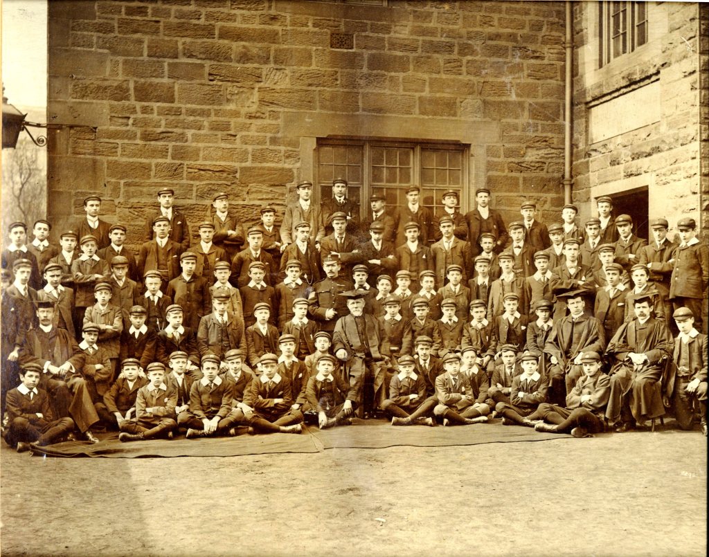 Black and white photo of a large group of boys and men, presumably students and staff, posed outside a school building, likely Holgate Grammar School.