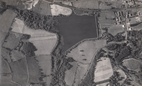 Aerial black and white photo of a landscape featuring large fields bordered by trees. A main road runs along the top, with several smaller roads on the right. Small clusters of buildings are visible in the upper right corner.