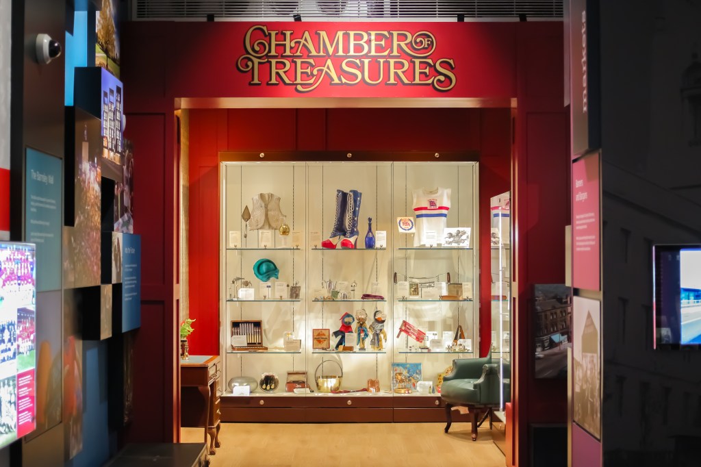 Display case labeled 'Chamber of Treasures' showcasing various artifacts and items on shelves, with a red background and green plant nearby.