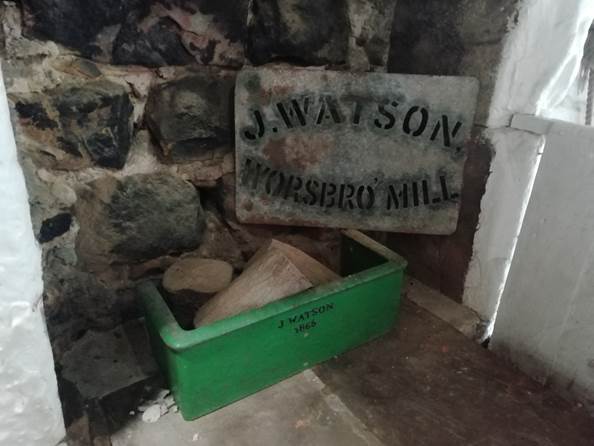 A vintage metal sign reading 'J.WATSON WORSBRO MILL' and a green wooden box labeled 'J WATSON 1865', placed against a stone wall with pieces of wood inside.
