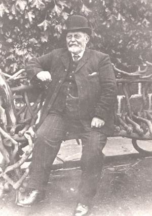 Portrait of an older man in a hat and suit, seated on a decorative wooden bench.