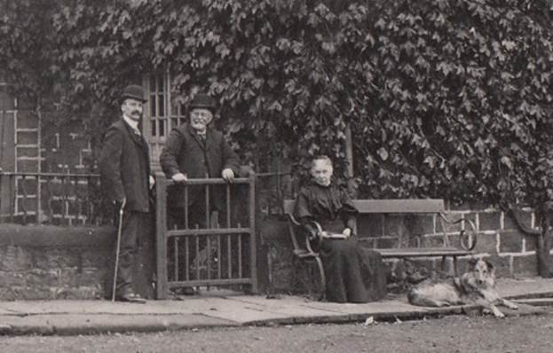 Historical photo of three individuals and a dog outside a building, with one man standing by a gate and another man and a woman seated nearby.