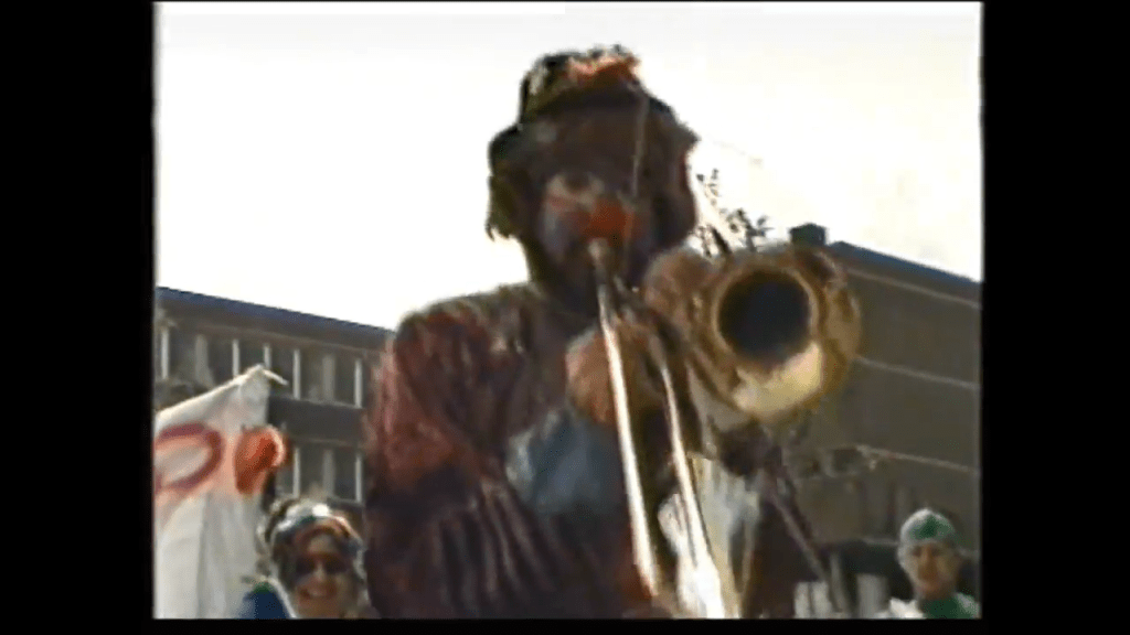 A performer in a colorful costume plays a trombone during a parade, surrounded by people celebrating.