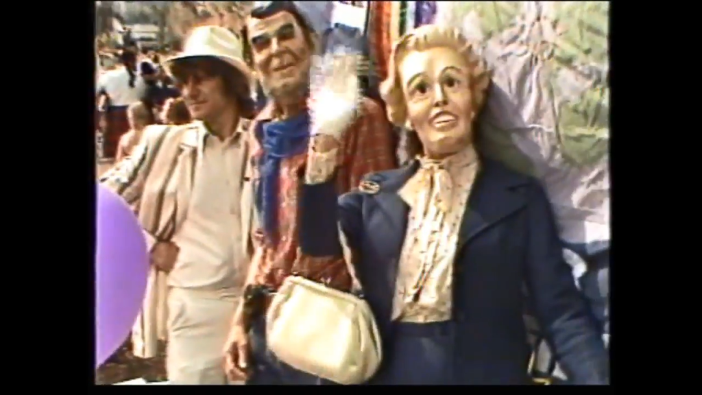 A group of people at a parade in the 1980s, with a man in a hat and a woman holding a puppet resembling a well-dressed lady, surrounded by colorful decorations.