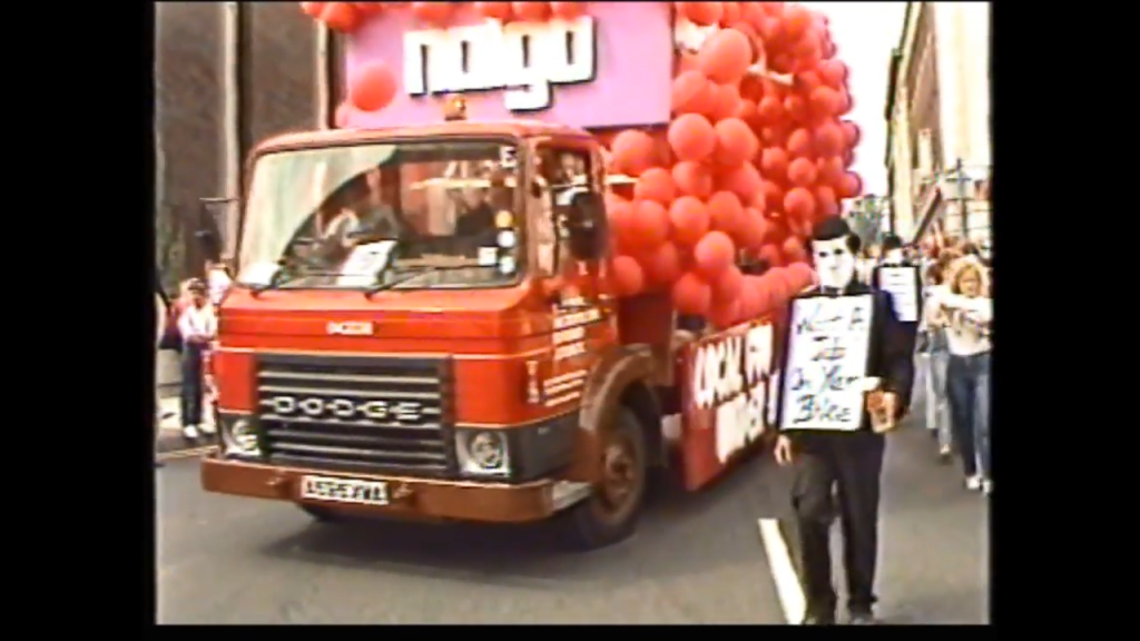 A red Dodge truck filled with red balloons drives in a parade, accompanied by a person in a suit and white mask holding a sign.