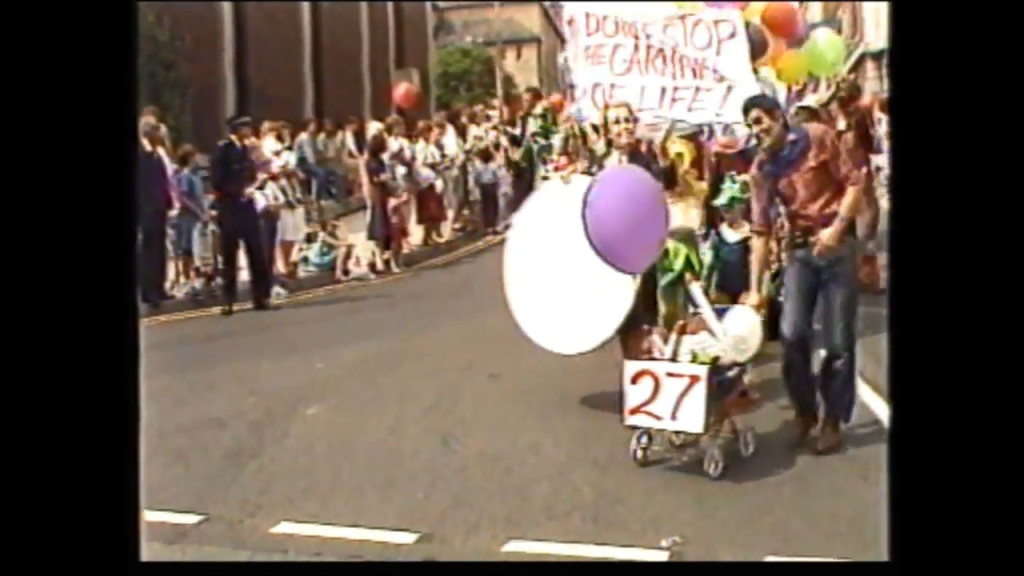 A lively parade scene featuring participants with balloons and a stroller numbered 27, as crowds watch along the street.