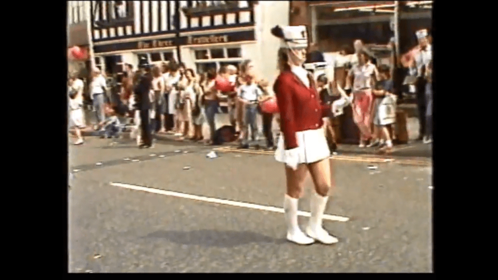 A parade scene featuring a woman in a red jacket and white skirt, standing on a street. She is holding a baton, while a crowd watches in the background.