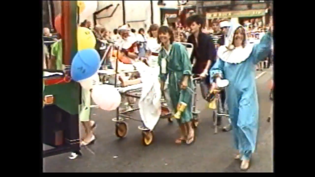Participants in a colorful parade, wearing themed costumes and pushing decorated carts, walk along a street filled with spectators, balloons in the background.