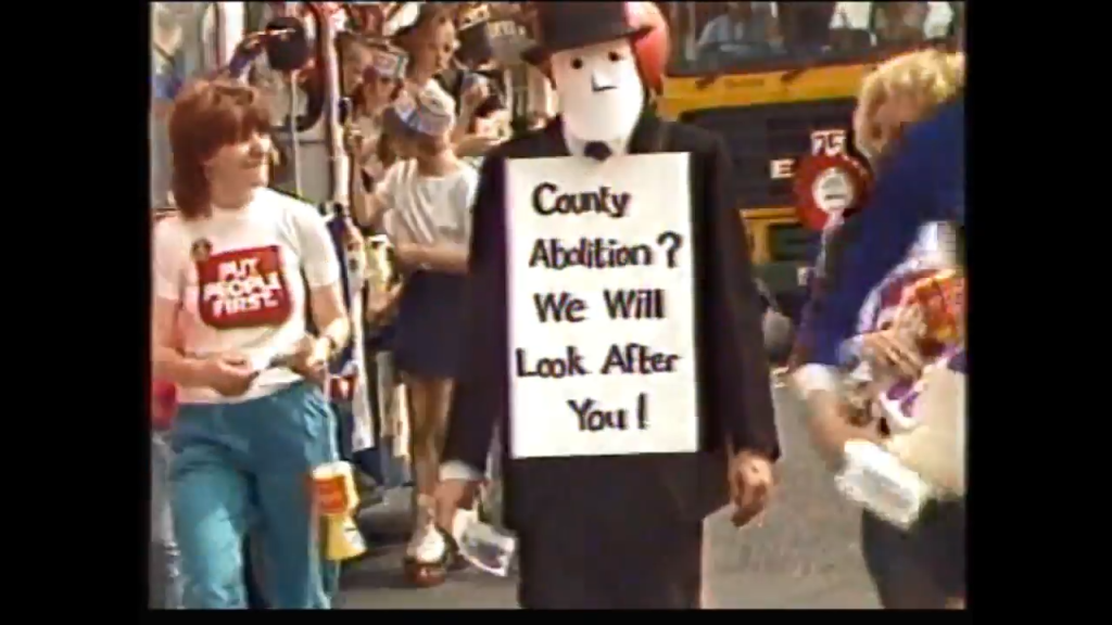 A person dressed in a suit with a mask holds a sign reading 'County Abolition? We Will Look After You!' while walking in a parade, accompanied by other parade participants.