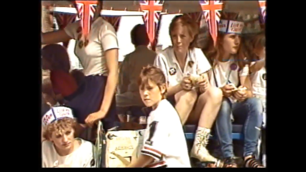 A group of children wearing white shirts and festive hats decorated with Union Jack patterns, sitting together during a parade, with decorative bunting in the background.