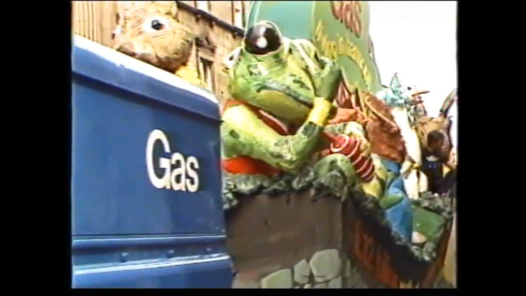 A colorful float from the Mayor's Parade featuring a frog and various creatures, with a blue vehicle labeled 'Gas' in the foreground.