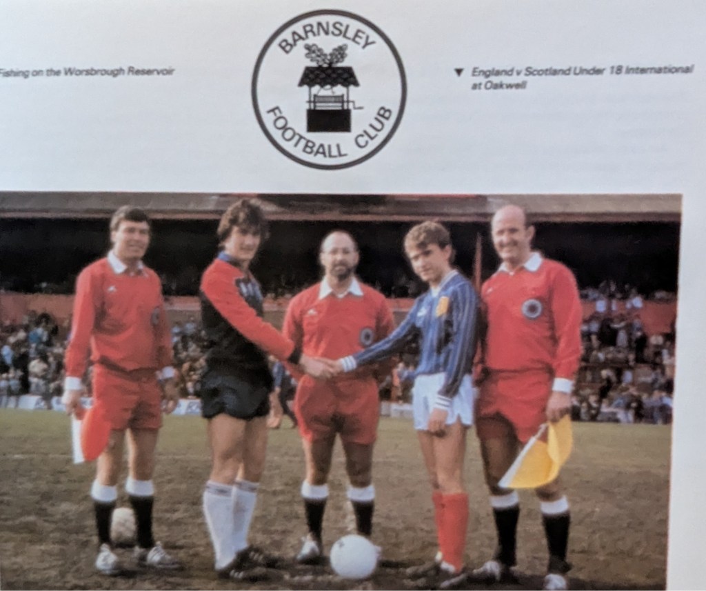 Five officials and players shake hands before the England vs Scotland Under 18 International match at Oakwell, with spectators in the background.
