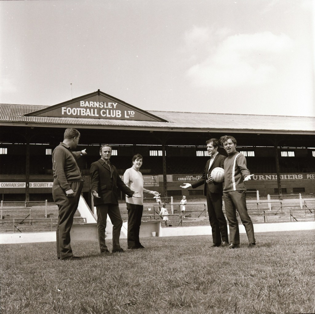 A group of five individuals standing on the pitch at Barnsley Football Club's stadium, with the club's sign visible in the background.
