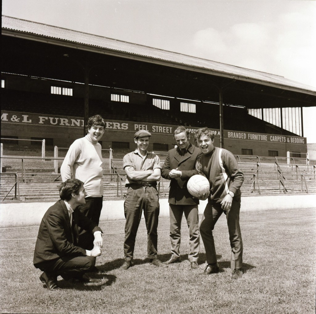 Black and white photograph of five men on a football field, posing with a soccer ball in front of a stadium with a sign for M&L Furnishers on Peel Street.