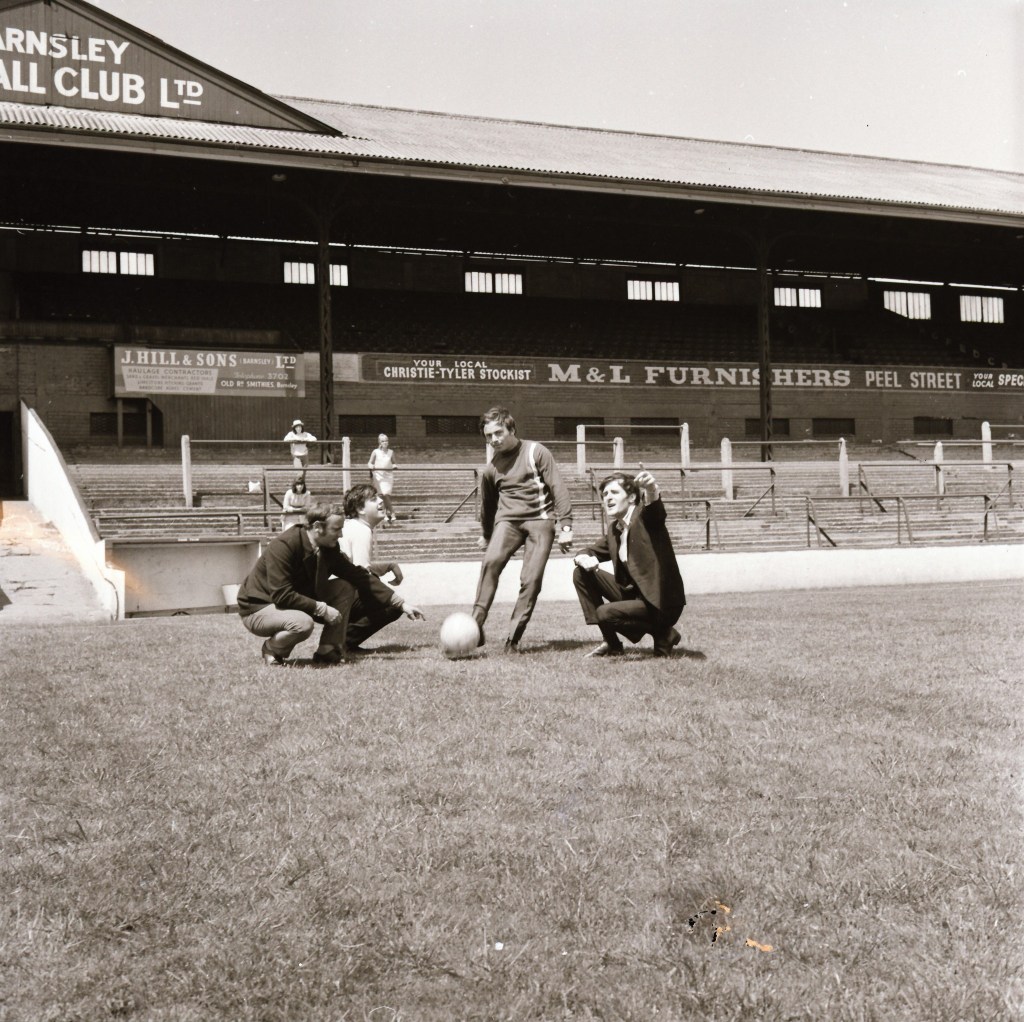 Three men gathered on the grass at Barnsley Football Club's stadium, with one man preparing to kick a football. The background features the stadium's grandstand and advertisements.