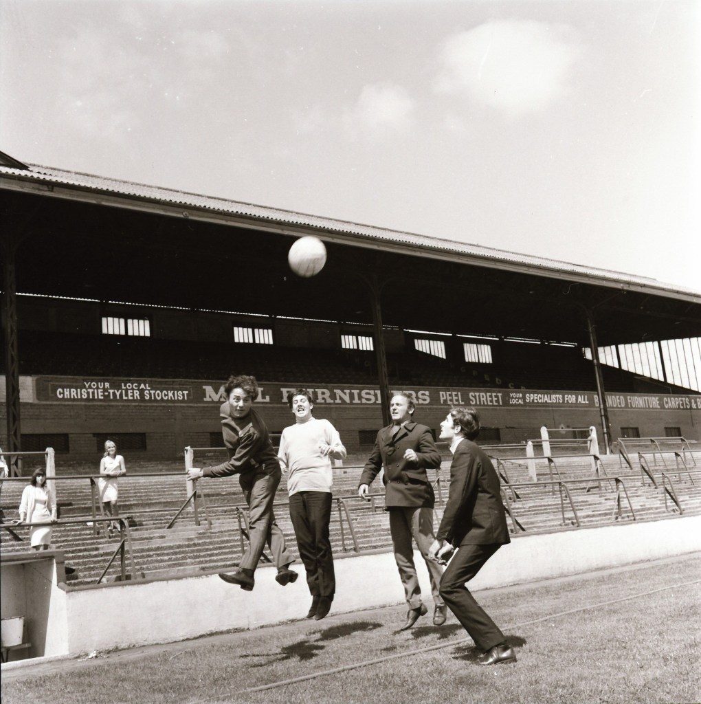 A black and white photograph of four men on a sports field, jumping to kick a football, with a stadium structure and a woman in the background.
