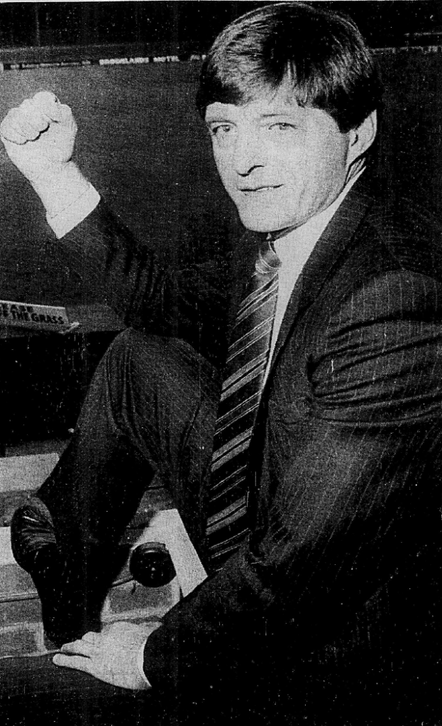 A black and white image of a man in a suit with a striped tie sitting on a stool, raising his fist in a triumphant gesture.