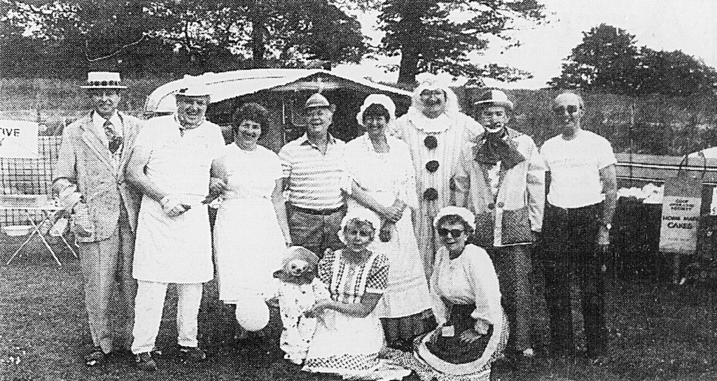 A black-and-white group photograph of individuals at an outdoor event, featuring people dressed in various attire, including a clown and others in white uniforms, smiling and posing in front of a tent.