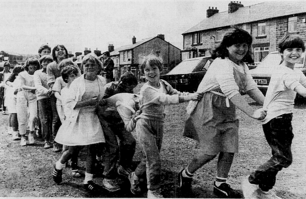 A group of children participates in a conga line on a street, smiling and enjoying themselves, with houses and cars visible in the background.