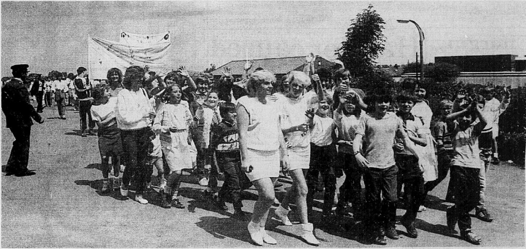A black and white photograph of a parade with children and adults walking together, some waving and holding banners. In the background, a police officer stands, and buildings are visible.