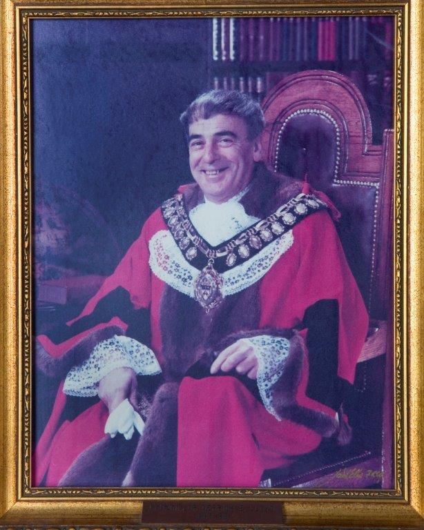 Portrait of Coun. Derrick Lloyd, the Mayor of Barnsley, in ceremonial robes, seated with a smile, surrounded by a library backdrop.