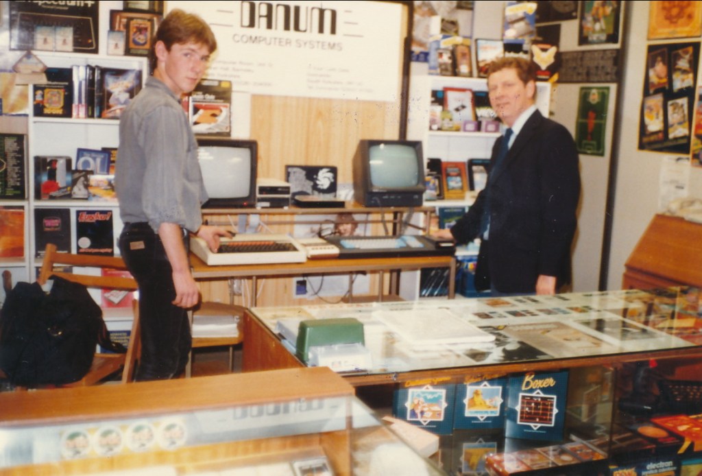 A young man and an older man stand in a computer store surrounded by various computer systems and memorabilia, with a computer monitor in the background.