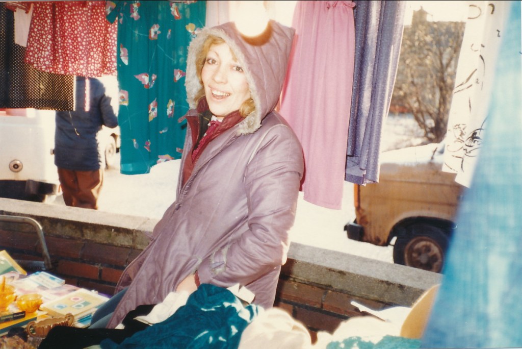 A smiling market trader wearing a pink coat stands near clothes hanging on a line, with what appears to be a vintage vehicle in the background.