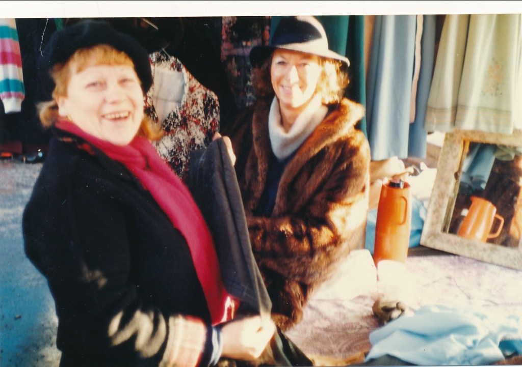 Two women smiling and interacting while surrounded by clothing and other items at an outdoor market.