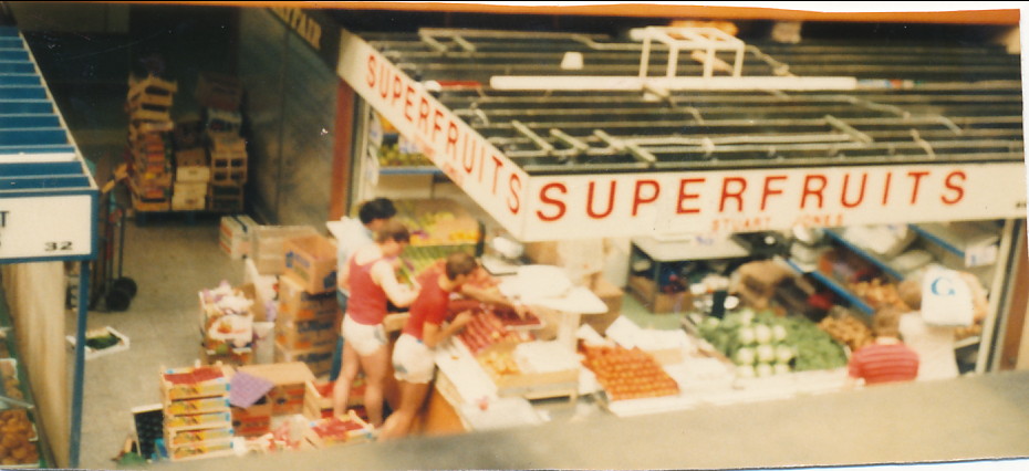 An overhead view of a fruit stall named 'Superfruits' in a market, with several people arranging produce and customers browsing.